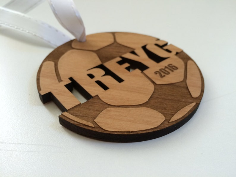 May include: A wooden soccer ball ornament with the name "TRAYCE" and the year "2016" engraved on it. The ornament is brown and has a white ribbon attached to it.