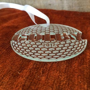 Pu&ograve; includere: Un ornamento di vetro trasparente con un motivo a pallina da golf e la parola "GOLLEY" e l'anno "2017" incisi sulla superficie.