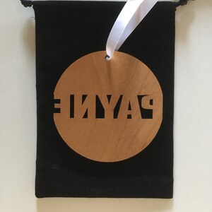 May include: A round wooden ornament with the word "PLAY" cut out in black lettering. The ornament is hanging from a white ribbon and is in a black velvet bag.