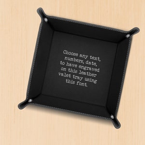 May include: Black leather valet tray with white stitching and rounded corners. The tray has text that reads, "Choose any text, numbers, date, to have engraved on this leather valet tray using this font."