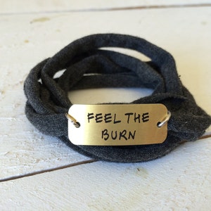 Puede incluir: Un brazalete de tela gris con una etiqueta de metal dorado que dice "Feel the Burn".