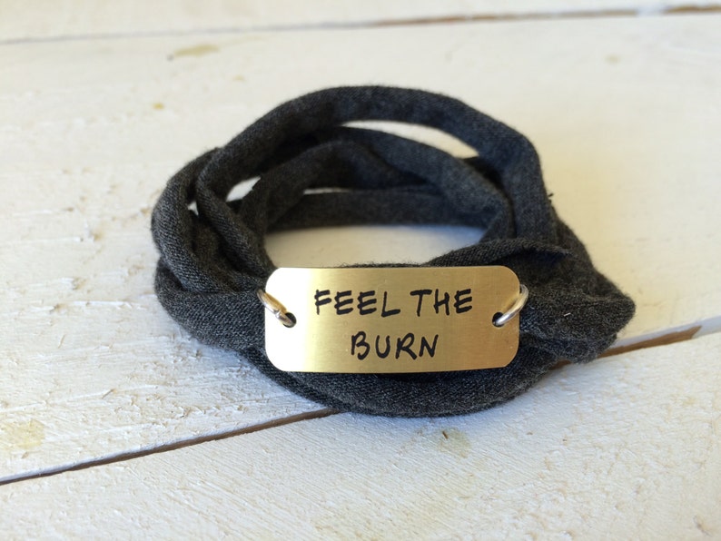 Puede incluir: Un brazalete de tela gris con una etiqueta de metal dorado que dice "Feel the Burn".