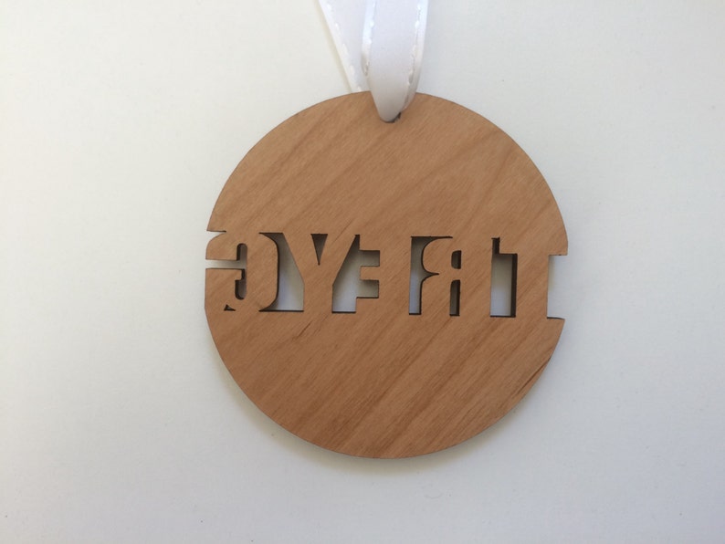 May include: A round, wooden ornament with the word "First" cut out in the center. The ornament is hanging from a white ribbon.