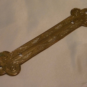 Op de afbeelding: Een decoratief stuk van verguld metaal met een bladontwerp en twee gaten. Het stuk heeft een lange, rechthoekige vorm met afgeronde uiteinden.