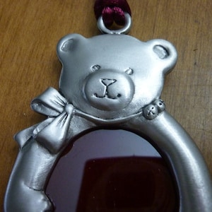Puede incluir: Un adorno de oso de peluche de peltre plateado con un óvalo de vidrio rojo en el centro. El oso tiene un lazo en el brazo y una pequeña campana en la oreja.