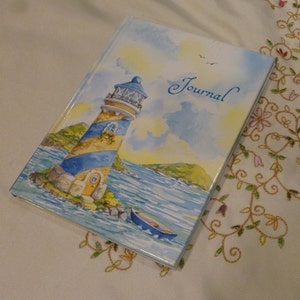 May include: A blue and yellow lighthouse with a white roof and a blue door on a watercolor background. The word "Journal" is written in cursive on the cover.