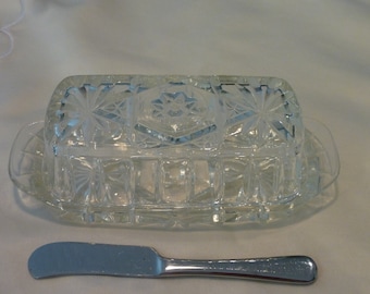 Op de afbeelding: Een glazen botervloot met een deksel en een bijpassend mes. De schaal heeft een geometrisch patroon dat in het glas is geëtst.