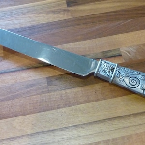 Puede incluir: Un cuchillo para pastel de plata con un mango decorativo con un diseño de vid y espiral. El cuchillo está sobre una superficie de madera.