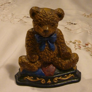 Könnte beinhalten: Ein Türstopper aus Gusseisen in Form eines Teddybären mit einer blauen Schleife. Der Bär sitzt auf einem bunten, gemusterten Sockel.
