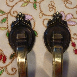 May include: A pair of antique brass drawer pulls with an ornate design. The pulls feature a dark, decorative backplate and a curved, gold-toned handle. Suitable for a cabinet or dresser.