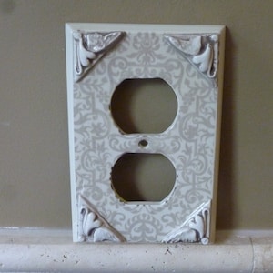 Pode incluir: Tampa de tomada de cerâmica branca com um padrão damasco decorativo cinza e branco. A tampa tem uma tomada dupla e é projetada para se ajustar a uma tomada elétrica padrão.