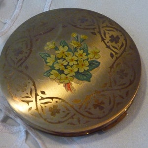 Op de afbeelding: Een goudkleurig compact met een bloemenontwerp. Het compact heeft een boeket gele bloemen met groene bladeren op een gouden achtergrond. Het compact heeft een decoratieve rand met een herhalend patroon.