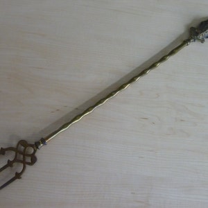 Könnte beinhalten: Eine lange, goldfarbene Gabel aus Metall mit einem gedrehten Griff und einem dekorativen Kopf. Die Gabel hat drei Zinken und ist zum Servieren von Speisen gedacht.