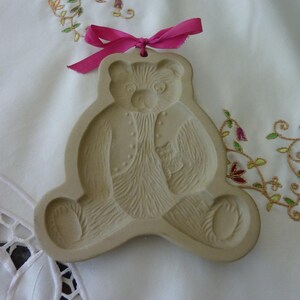 May include: A beige ceramic cookie cutter shaped like a teddy bear wearing a jacket. The cutter has a pink ribbon attached to the top.
