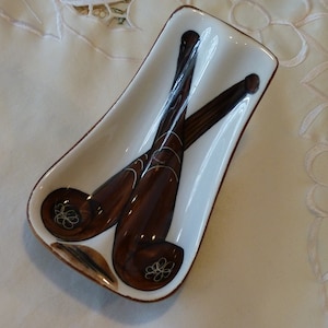 May include: White ceramic spoon rest with a brown and black painted design of two crossed spoons. The spoons have white floral details.