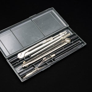 Könnte beinhalten: Eine graue Kunststoffbox mit einem Satz silberner Zeichenwerkzeuge, darunter Zirkel und Teiler. Die Werkzeuge sind ordentlich in der geöffneten Box vor einem schwarzen Hintergrund angeordnet. Die Box ist rechteckig mit einem Klappdeckel.