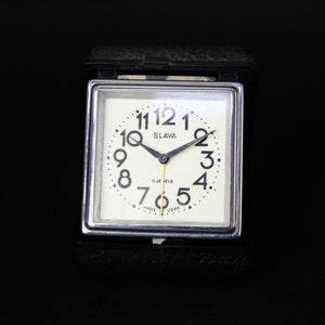 May include: A vintage square travel alarm clock with a cream-colored face, black numerals, and black hands. The clock face reads "SLAVA" and "17 JEWELS MADE USSR". The clock is encased in a silver frame and a black leatherette case.