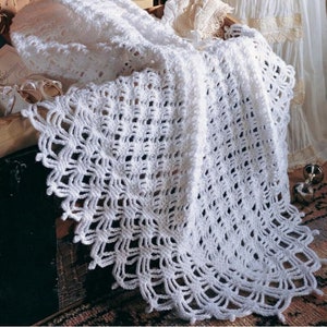 May include: A white, handmade crochet blanket with a delicate, openwork design. The blanket features a scalloped edge and is draped over a wooden surface. The intricate pattern and soft texture suggest a cozy, handcrafted item, perfect for a baby or as a decorative piece.