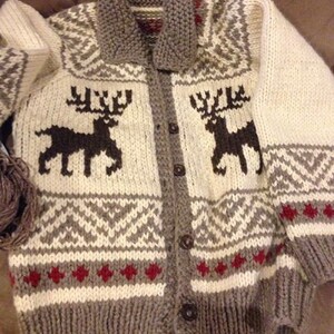 May include: A chunky knit cardigan featuring a cream-colored base with dark brown reindeer silhouettes on the chest. The garment includes a grey ribbed collar, cuffs, and hem, accented by geometric patterns in grey and small red diamond shapes.