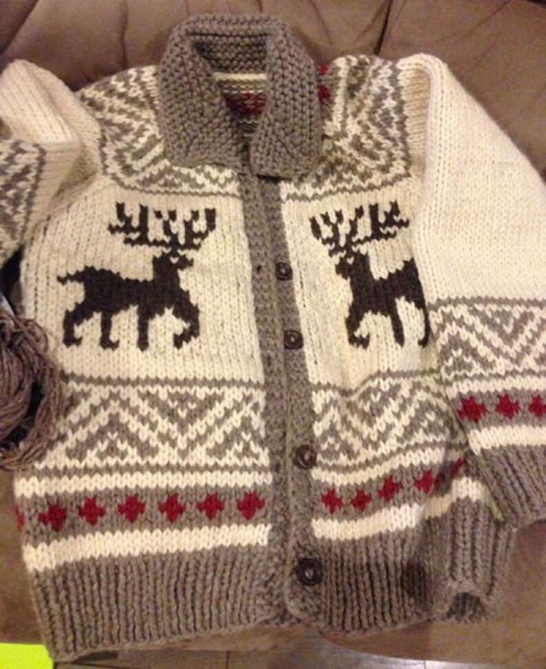 May include: A chunky knit cardigan featuring a cream-colored base with dark brown reindeer silhouettes on the chest. The garment includes a grey ribbed collar, cuffs, and hem, accented by geometric patterns in grey and small red diamond shapes.