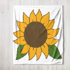 Op de afbeelding: Een wit kussen met een geel zonnebloemontwerp. De zonnebloem heeft een bruin centrum en groene bladeren.