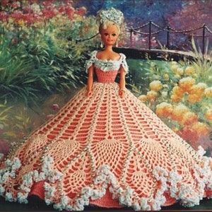 May include: A crocheted doll dress in a peach color with white lace trim and pearl-like beads. The dress has a full skirt and is designed for a doll with a waist measurement of approximately 11 inches.