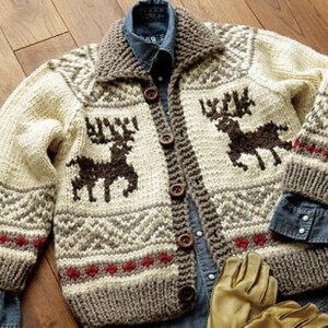 May include: A chunky knit cardigan featuring a cream base with brown reindeer motifs, a large collar, and five dark brown buttons. The sweater displays geometric patterns in brown and red accents along the hem and sleeves, layered over a blue denim shirt.