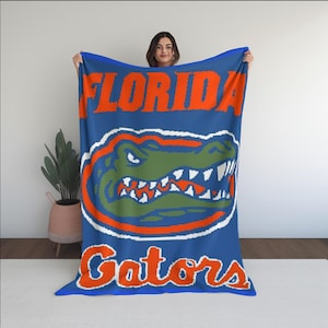 Puede incluir: Manta azul con las palabras "FLORIDA" y "Gators" en naranja. La manta presenta un logotipo de caimán verde y naranja con detalles en blanco. La manta está sostenida por una persona.