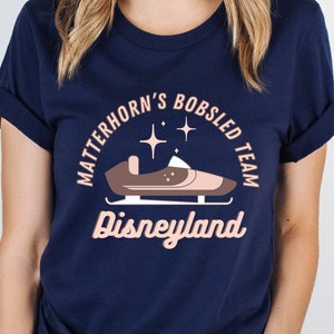 May include: A navy blue t-shirt with a pink graphic of a bobsled on the Matterhorn ride at Disneyland. The text "Matterhorn's Bobsled Team Disneyland" is printed on the shirt.