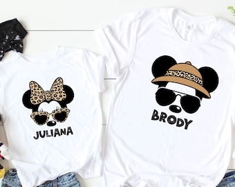 Op de afbeelding: Twee witte T-shirts met een cartoon afbeelding van Mickey Mouse. Het shirt van het meisje heeft een strik op Mickey's hoofd en zegt "Juliana". Het shirt van de jongen heeft Mickey met een safarihoed en zonnebril en zegt "Brody".