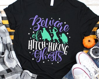 May include: Black t-shirt with a purple and green design that says "Beware of Hitchhiking Ghosts". The design features silhouettes of the three ghosts from the Haunted Mansion attraction at Disney.