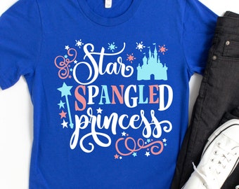 Può includere: Una maglietta blu reale con il testo "Star Spangled Princess" in bianco con accenti rosa e blu. Il design include anche un castello bianco e stelle.