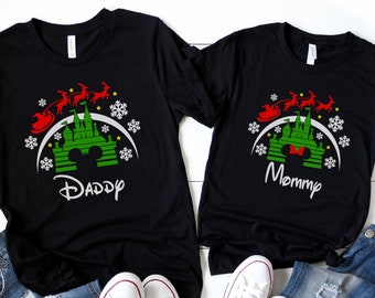 Op de afbeelding: Zwarte T-shirts met een groen en rood kerstthema met een kasteel en de kerstman die eroverheen vliegt. De tekst "Daddy" en "Mommy" staat op de shirts gedrukt.