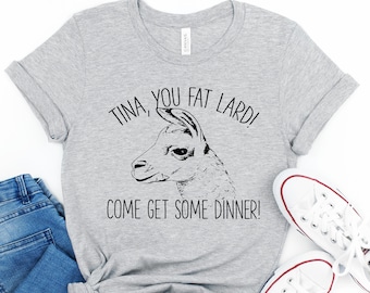 May include: A gray t-shirt with a black and white illustration of a llama and the text "TINA, YOU FAT LARD! COME GET SOME DINNER!"