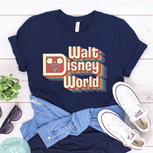 Puede incluir: Camiseta azul marino con un dise&ntilde;o retro de "Walt Disney World" con letras coloridas en capas y el logotipo de Mickey Mouse. La imagen tambi&eacute;n muestra zapatillas blancas y una camisa vaquera.