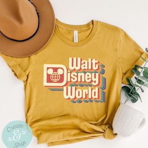 Puede incluir: Camiseta de color amarillo mostaza con la frase "Walt Disney World" en una fuente retro. El dise&ntilde;o incluye un logotipo de la cabeza de Mickey Mouse y rayas coloridas. Un sombrero marr&oacute;n y una taza blanca est&aacute;n en el fondo.