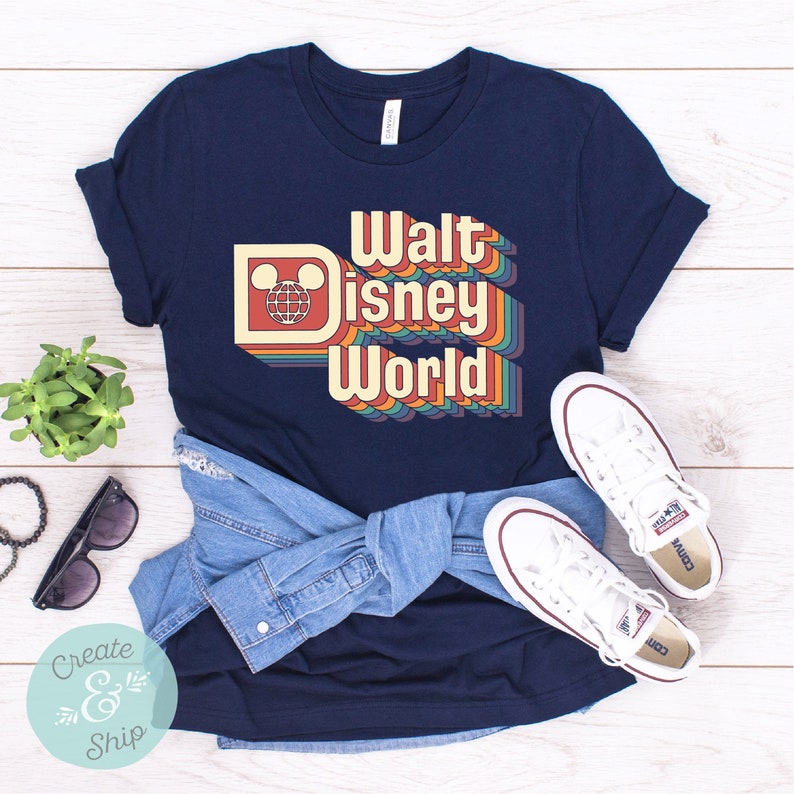Puede incluir: Camiseta azul marino con un dise&ntilde;o retro de "Walt Disney World" con letras coloridas en capas y el logotipo de Mickey Mouse. La imagen tambi&eacute;n muestra zapatillas blancas y una camisa vaquera.