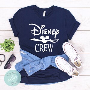 以下が含まれることがあります： 白い文字で「Disney Crew」と書いてある紺色のTシャツ。白いミッキーマウスの頭が耳をつけていて、白い剣の上に載っています。