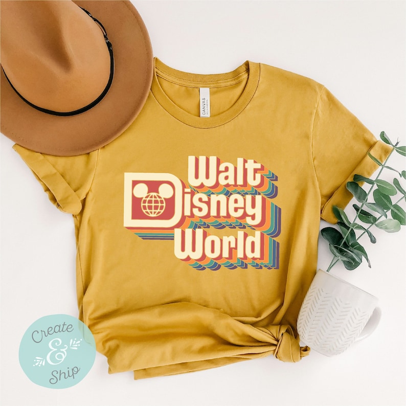 Puede incluir: Camiseta de color amarillo mostaza con la frase "Walt Disney World" en una fuente retro. El dise&ntilde;o incluye un logotipo de la cabeza de Mickey Mouse y rayas coloridas. Un sombrero marr&oacute;n y una taza blanca est&aacute;n en el fondo.