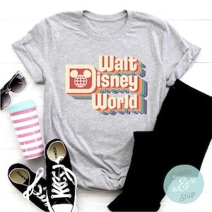 Puede incluir: Una camiseta gris con las palabras "Walt Disney World" en un dise&ntilde;o retro arco&iacute;ris. La camiseta presenta un logotipo de la cabeza de Mickey Mouse. Pantalones negros, zapatillas Converse negras, gafas de sol y un vaso rosa y azul tambi&eacute;n est&aacute;n en la imagen.