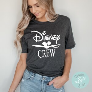 以下が含まれることがあります： 「Disney Crew」と白で書かれたグラフィックデザインが施された濃いグレーのTシャツ。Crewの文字の上にミッキーマウスの頭が描かれている。