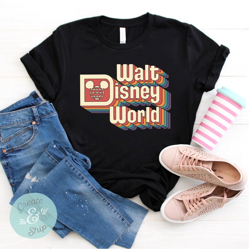 Puede incluir: Camiseta negra con un dise&ntilde;o retro con las palabras "Walt Disney World" en una fuente colorida en capas. El dise&ntilde;o incluye una cabeza de Mickey Mouse y un icono de globo. La camiseta se combina con vaqueros azules y zapatillas rosas.