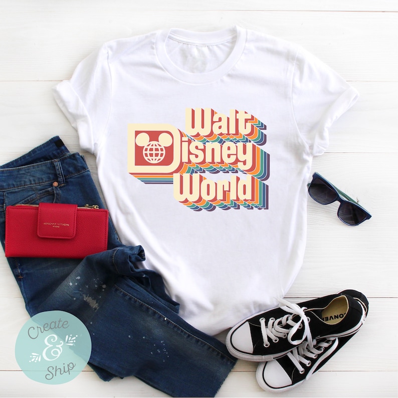 Puede incluir: Camiseta blanca con un dise&ntilde;o retro de "Walt Disney World" en colores arco&iacute;ris. El dise&ntilde;o incluye el ic&oacute;nico logotipo de la cabeza de Mickey Mouse y el globo terr&aacute;queo. La camiseta se combina con vaqueros azules, una cartera roja, gafas de sol y zapatillas negras.