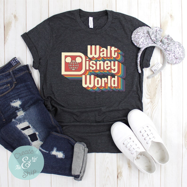 Puede incluir: Camiseta gris oscuro con la frase "Walt Disney World" en un dise&ntilde;o retro arco&iacute;ris. La camiseta se combina con vaqueros azules desgastados, zapatillas blancas y orejas de rat&oacute;n con lentejuelas. Un atuendo informal.