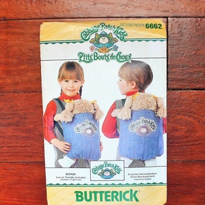 May include: A Butterick sewing pattern envelope for Cabbage Patch Kids, numbered 6662. The front features an illustration of a child wearing a blue denim-style doll carrier with a doll inside. The text includes Cabbage Patch Kids, P'tits Bouts de Choux, and Butterick.