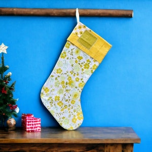 May include: A yellow and white floral Christmas stocking hangs from a wooden rod against a blue wall. A small artificial Christmas tree with gold ornaments sits on a wooden table in front of the stocking.