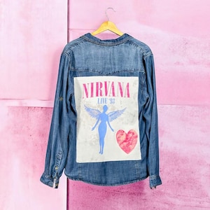 May include: A denim shirt with long sleeves, hanging on a wooden hanger. The back of the shirt features a white rectangle with the text "NIRVANA LIVE '93" in pink. Below the text is a blue angel figure with wings and a pink heart.