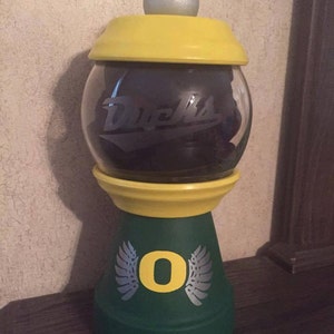 May include: A green and yellow ceramic candy dispenser with a silver lid and a clear glass jar. The jar has the word "Ducks" written on it. The base of the dispenser has a silver "O" with wings on either side.