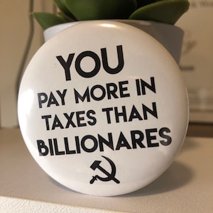 May include: White button with black text that reads "YOU PAY MORE IN TAXES THAN BILLIONAIRES" with a black hammer and sickle symbol below the text.