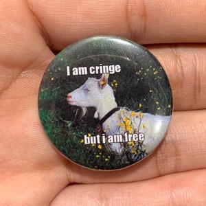 Könnte beinhalten: Ein runder Knopf mit einem Bild einer weißen Ziege mit einem schwarzen Halsband, die in einem Feld aus grünem Gras und gelben Blumen liegt. Die Ziege hat die Augen geschlossen und den Kopf zur Seite geneigt. Der Text "I am cringe but I am free" ist auf dem Knopf gedruckt.